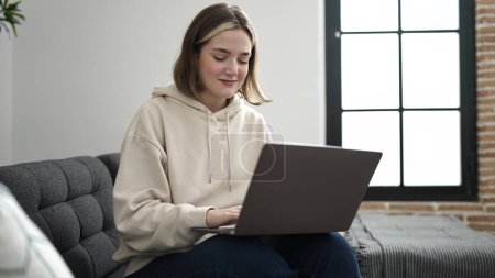 Foto de Mujer rubia joven usando el ordenador portátil sentado en el sofá en casa - Imagen libre de derechos