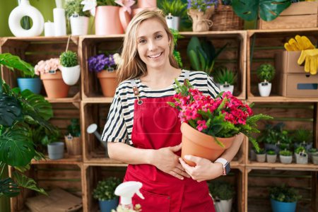 Foto de Joven mujer caucásica que trabaja en floristería sosteniendo planta con aspecto positivo y feliz de pie y sonriendo con una sonrisa confiada mostrando los dientes - Imagen libre de derechos