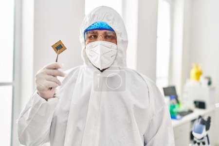 Téléchargez les photos : Jeune homme latin scientifique portant uniforme de protection covid tenant puce processeur cpu au laboratoire - en image libre de droit