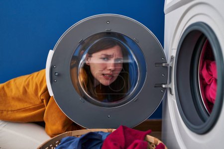 Foto de Mujer morena joven mirando a través de la ventana de la lavadora sin pistas y expresión confusa. concepto de duda. - Imagen libre de derechos