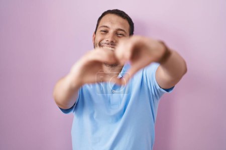 Foto de Hombre hispano de pie sobre fondo rosa sonriendo en el amor haciendo forma de símbolo del corazón con las manos. concepto romántico. - Imagen libre de derechos