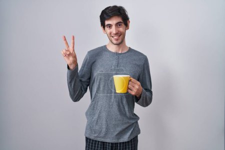 Téléchargez les photos : Jeune homme hispanique portant un pyjama buvant une tasse de café souriant avec le visage heureux clin d'oeil à la caméra faisant signe de victoire avec les doigts. numéro deux. - en image libre de droit