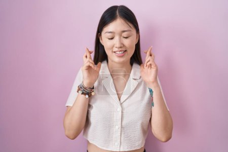 Téléchargez les photos : Jeune femme chinoise debout sur fond rose geste doigt croisé souriant avec espoir et les yeux fermés. chance et concept superstitieux. - en image libre de droit