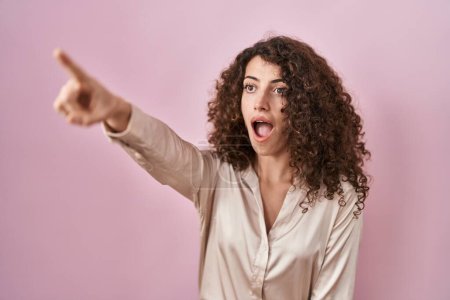 Foto de Mujer hispana con el pelo rizado de pie sobre fondo rosa apuntando con el dedo sorprendido delante, boca abierta expresión asombrada, algo en el frente - Imagen libre de derechos