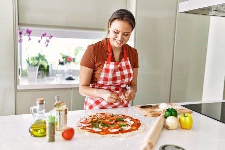 Photo for Young beautiful hispanic woman smiling confident putting onion on pizza at the kitchen - Royalty Free Image