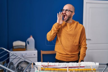 Photo for Young bald man with beard hanging clothes at clothesline clueless and confused with open arms, no idea and doubtful face. - Royalty Free Image