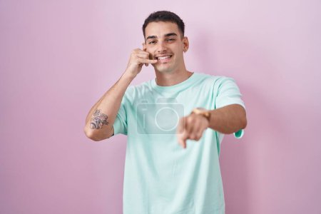 Téléchargez les photos : Bel homme hispanique debout sur fond rose souriant faire parler au téléphone geste et vous pointant du doigt. Appelle-moi. - en image libre de droit