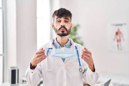 Photo for Young hispanic doctor man with beard holding safety mask looking at the camera blowing a kiss being lovely and sexy. love expression. - Royalty Free Image