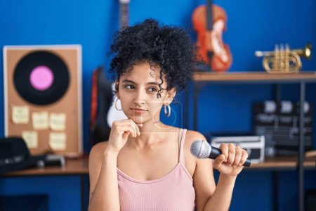 Photo for Hispanic woman with curly hair singing song using microphone at music studio serious face thinking about question with hand on chin, thoughtful about confusing idea - Royalty Free Image