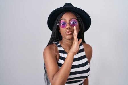 Foto de Joven afroamericano con trenzas con sombrero y gafas de sol mano en boca contando rumores secretos, susurrando conversación maliciosa - Imagen libre de derechos