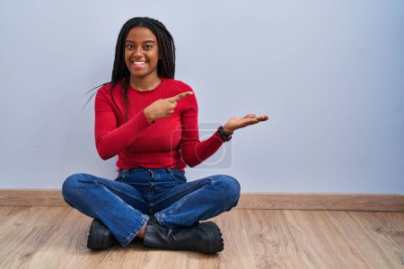Téléchargez les photos : Jeune afro-américain avec des tresses assis sur le sol à la maison étonné et souriant à la caméra tout en présentant avec la main et pointant du doigt. - en image libre de droit
