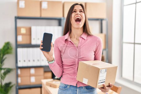 Photo for Young hispanic woman working at small business ecommerce showing smartphone screen angry and mad screaming frustrated and furious, shouting with anger looking up. - Royalty Free Image