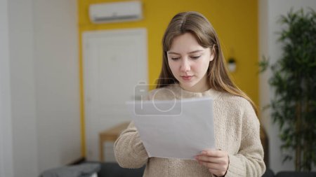 Foto de Joven mujer rubia leyendo documento de pie en casa - Imagen libre de derechos