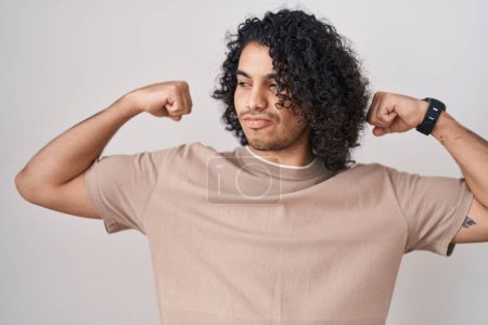 Téléchargez les photos : Homme hispanique avec les cheveux bouclés debout sur fond blanc montrant les muscles des bras souriant fier. concept de fitness. - en image libre de droit