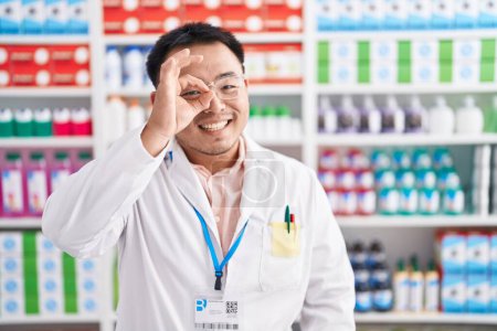 Téléchargez les photos : Jeune homme chinois travaillant à la pharmacie pharmacie faisant un geste correct avec le sourire de la main, les yeux regardant à travers les doigts avec un visage heureux. - en image libre de droit