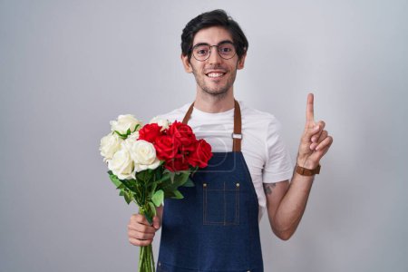 Foto de Joven hombre hispano sosteniendo ramo de rosas blancas y rojas sonriendo asombrado y sorprendido y señalando con los dedos y los brazos levantados. - Imagen libre de derechos