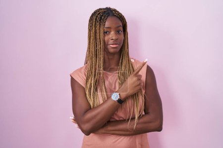 Téléchargez les photos : Femme afro-américaine avec des cheveux tressés debout sur fond rose pointant avec le doigt de la main sur le côté montrant la publicité, visage sérieux et calme - en image libre de droit
