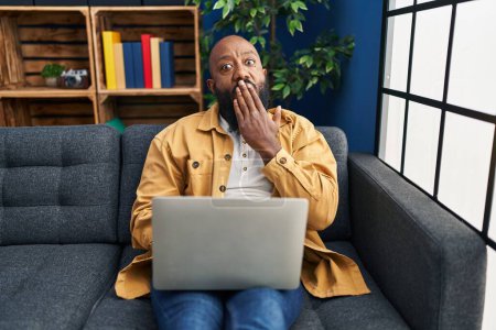 Téléchargez les photos : Afro-Américain utilisant un ordinateur portable à la maison assis sur le canapé couvrant la bouche avec la main, choqué et effrayé par l'erreur. expression surprise - en image libre de droit