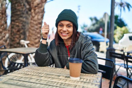 Foto de Mujer hispana joven bebiendo una taza de café al aire libre sonriendo feliz y positivo, pulgar hacia arriba haciendo excelente y signo de aprobación - Imagen libre de derechos
