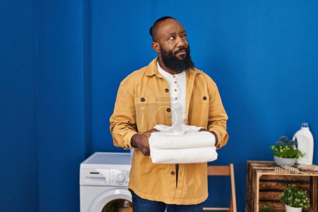 Téléchargez les photos : Afro-Américain tenant des serviettes propres à la buanderie souriant regardant vers le côté et regardant loin en pensant. - en image libre de droit