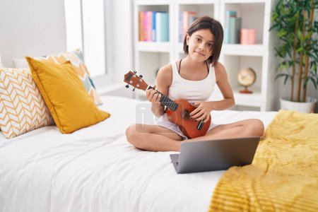 Téléchargez les photos : Adorable hispanic girl having online ukulele lesson sitting on bed at bedroom - en image libre de droit