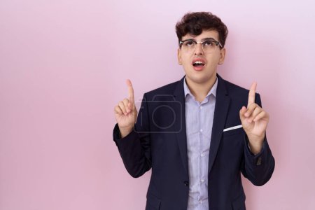 Photo for Young non binary man with beard wearing suit and tie amazed and surprised looking up and pointing with fingers and raised arms. - Royalty Free Image