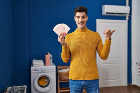 Foto de Joven hombre hispano en la lavandería sosteniendo shekels apuntando pulgar hacia el lado sonriendo feliz con la boca abierta - Imagen libre de derechos