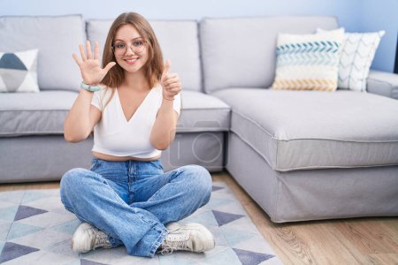 Téléchargez les photos : Jeune femme caucasienne assise sur le sol au salon montrant et pointant vers le haut avec les doigts numéro six tout en souriant confiant et heureux. - en image libre de droit