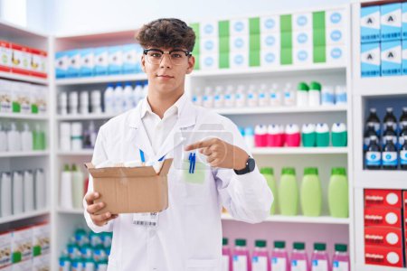 Téléchargez les photos : Adolescent hispanique travaillant à la pharmacie drugstore tenant boîte avec des pilules souriant heureux pointant avec la main et le doigt - en image libre de droit