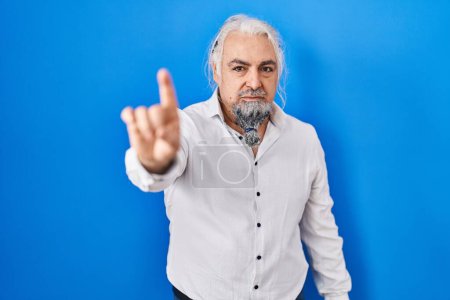 Foto de Hombre de mediana edad con el pelo gris de pie sobre fondo azul apuntando con el dedo hacia arriba y la expresión enojada, que no muestra ningún gesto - Imagen libre de derechos
