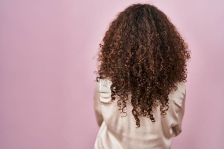 Foto de Mujer hispana con el pelo rizado de pie sobre fondo rosa de pie hacia atrás mirando hacia otro lado con los brazos cruzados - Imagen libre de derechos