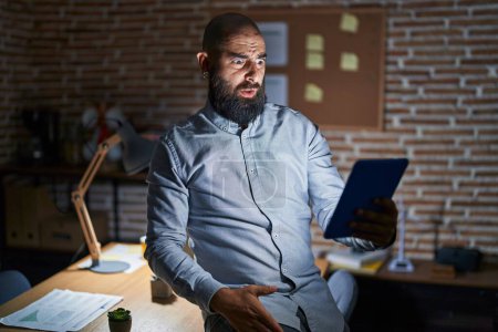 Photo for Young hispanic man with beard and tattoos working at the office at night in shock face, looking skeptical and sarcastic, surprised with open mouth - Royalty Free Image