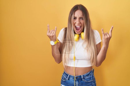 Téléchargez les photos : Jeune femme blonde debout sur fond jaune portant des écouteurs criant avec une expression folle faisant symbole rock avec les mains en l'air. star de la musique. concept de musique lourde. - en image libre de droit