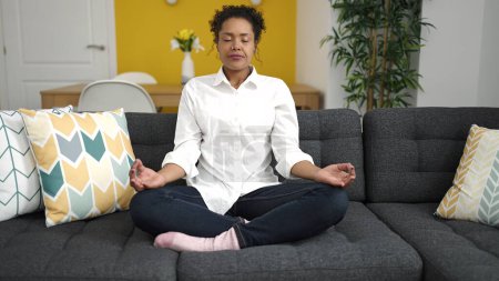 Téléchargez les photos : Femme afro-américaine faisant de l'exercice de yoga assis sur le canapé à la maison - en image libre de droit