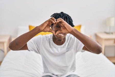 Foto de Hombre afroamericano haciendo gesto de corazón sentado en la cama en el dormitorio - Imagen libre de derechos
