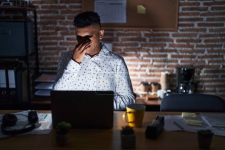 Photo for Young hispanic man working at the office at night smelling something stinky and disgusting, intolerable smell, holding breath with fingers on nose. bad smell - Royalty Free Image