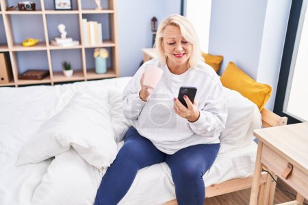 Foto de Middle age blonde woman using smartphone drinking coffee sitting on bed at bedroom - Imagen libre de derechos