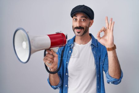 Foto de Hombre hispano con barba gritando a través de megáfono haciendo signo bien con los dedos, sonriendo gesto amistoso excelente símbolo - Imagen libre de derechos