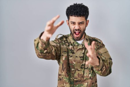 Photo for Arab man wearing camouflage army uniform shouting frustrated with rage, hands trying to strangle, yelling mad - Royalty Free Image
