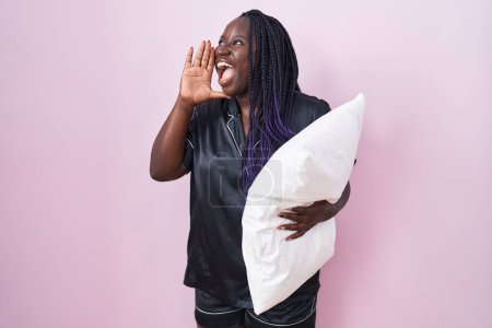 Foto de Mujer africana joven con pijama abrazando almohada gritando y gritando fuerte a un lado con la mano en la boca. concepto de comunicación. - Imagen libre de derechos