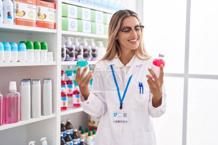 Foto de Joven mujer rubia farmacéutica sonriendo confiada sosteniendo botellas de medicamentos en la farmacia - Imagen libre de derechos
