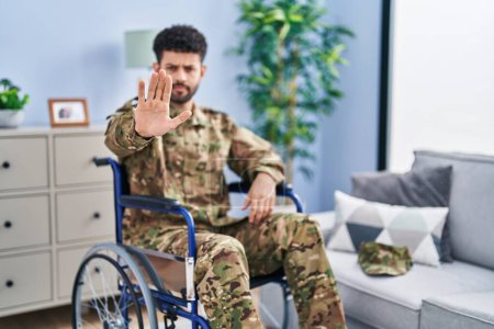 Photo for Arab man wearing camouflage army uniform sitting on wheelchair doing stop sing with palm of the hand. warning expression with negative and serious gesture on the face. - Royalty Free Image