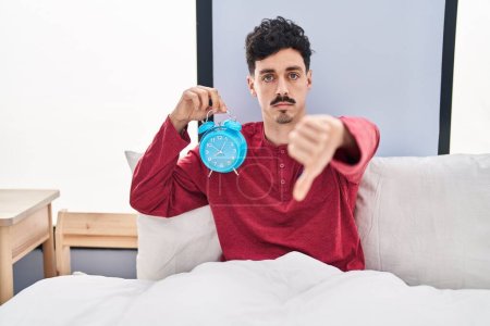 Photo for Hispanic man holding alarm clock in the bed with angry face, negative sign showing dislike with thumbs down, rejection concept - Royalty Free Image