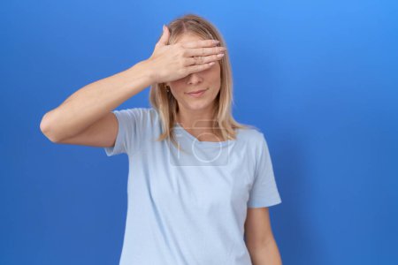 Foto de Mujer caucásica joven que usa una camiseta azul casual que cubre los ojos con la mano, luciendo seria y triste. concepto de ocultamiento y rechazo sin visión - Imagen libre de derechos