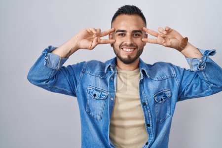 Foto de Joven hombre hispano de pie sobre un fondo aislado haciendo símbolo de paz con los dedos sobre la cara, sonriendo alegre mostrando la victoria - Imagen libre de derechos