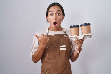 Foto de Mujer hispana joven con delantal de camarera profesional sosteniendo café sorprendido señalando con el dedo de la mano hacia un lado, boca abierta expresión asombrada. - Imagen libre de derechos