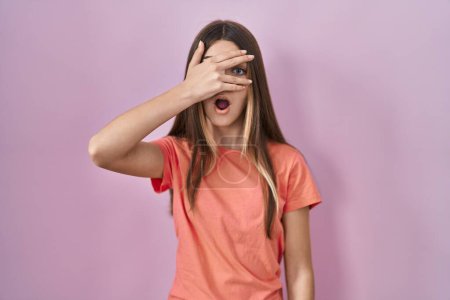Foto de Adolescente chica de pie sobre fondo rosa asomándose en shock cubriendo la cara y los ojos con la mano, mirando a través de los dedos con expresión avergonzada. - Imagen libre de derechos