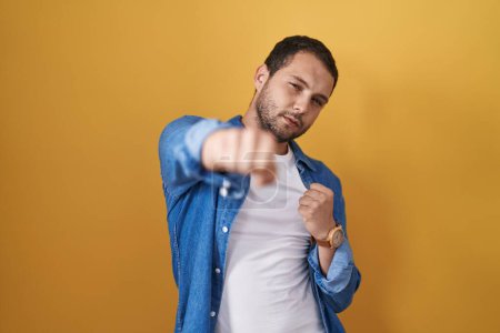 Photo for Hispanic man standing over yellow background punching fist to fight, aggressive and angry attack, threat and violence - Royalty Free Image