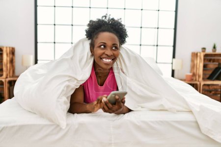 Téléchargez les photos : Femme afro-américaine utilisant un smartphone couché sur le lit dans la chambre - en image libre de droit