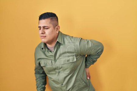Téléchargez les photos : Jeune homme hispanique debout sur fond jaune souffrant de maux de dos, touchant le dos avec la main, douleurs musculaires - en image libre de droit